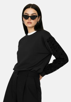 Kobieta w czarnej bluzie z koronkowymi rękawami, czarnych okularach przeciwsłonecznych typu cat-eye, srebrnych kolczykach kołach i czarnych spodniach z wysokim stanem na białym tle.