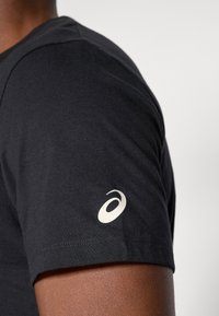 Közelkép egy fekete rövid ujjú pólót viselő személyről, amelyen fehér ASICS logó látható az ujján, egyszerű háttér előtt.