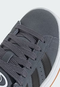 Zapatilla de ante gris con cordones planos grises trenzados, acentos de rayas negras y una suela de goma blanca con un patrón de relieve distintivo.
