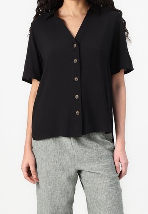 Kvinde iført en løs sort kortærmet skjorte med knapper kombineret med lysegrå teksturerede bukser mod en hvid baggrund.