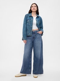 Világoskék farmer széles szárú nadrág magas derékkal, fehér crop top-pal és farmerdzsekivel párosítva. A modellen krém színű csíkos sportcipők vannak.