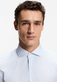 Młody mężczyzna o kręconych włosach, ubrany w białą koszulę z cienkimi niebieskimi pionowymi paskami, zwrócony przodem, o neutralnym wyrazie twarzy.