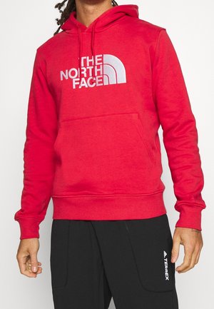 Rød hættetrøje lavet af blødt stof, med stort hvidt logo, frontlomme og justerbar hætte med snøre.