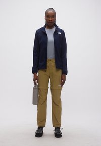 Γυναίκα όρθια, φοράει σκούρο μπλε μπουφάν, ανοιχτό γαλάζιο πουκάμισο, χακί παντελόνι με δυνατότητα μετατροπής, μαύρα παπούτσια πεζοπορίας, κρατώντας ένα γκρι παγούρι νερού.