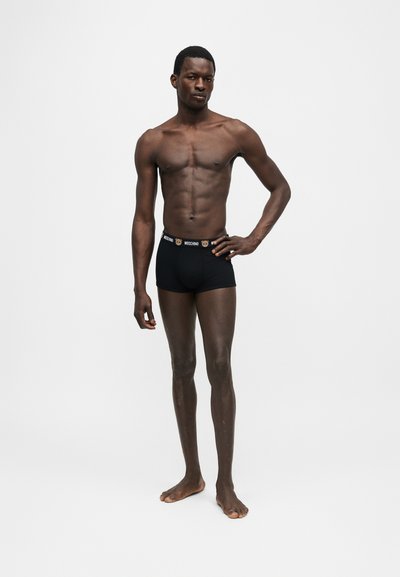 Αθλητικός άντρας όρθιος ξυπόλυτος, φοράει μαύρα μπόξερ Moschino με λογότυπο αρκουδάκι στη μέση, σε απλό λευκό φόντο.