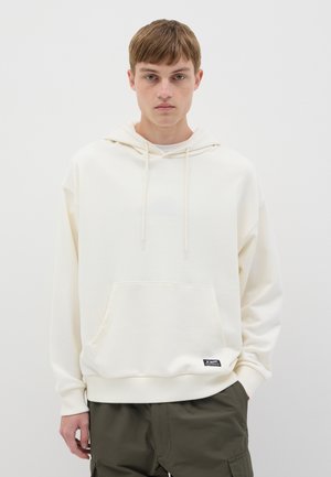 Sudadera crema clara con un bolsillo frontal, cordones en el cuello, puños y dobladillo acanalados, hecha de una tela suave, combinada con pantalones cargo oliva.