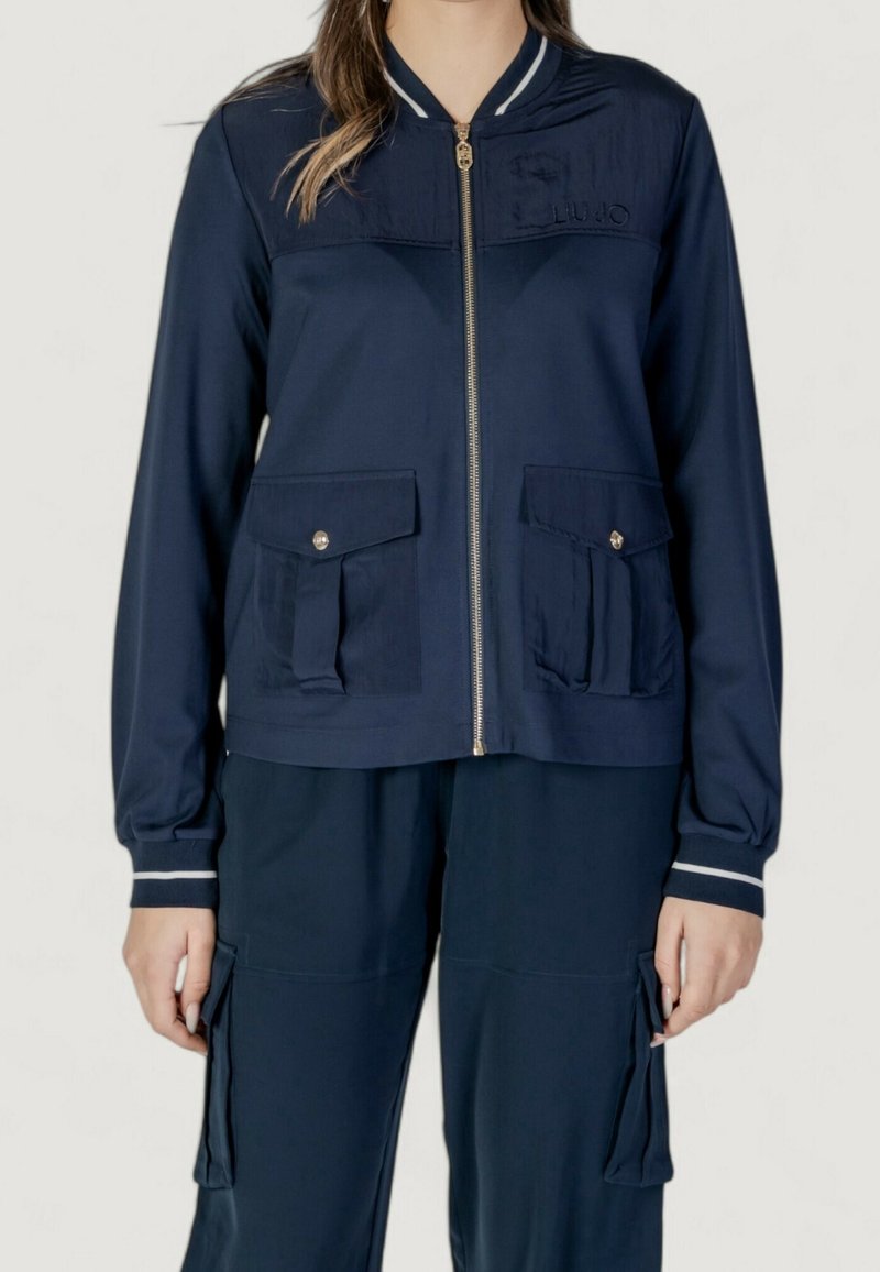 Femme portant une veste zippée bleu marine avec deux poches à rabat boutonnées et un pantalon cargo bleu marine assorti avec des poches latérales.