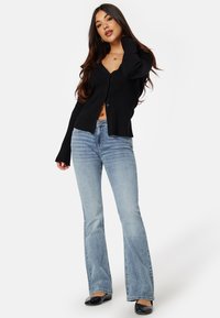 Schwarze gerippte Langarm-Cardigan mit Knöpfen vorne, kombiniert mit hellblauen Schlagjeans. Das Outfit hat eine verkürzte Silhouette und flache schwarze Schuhe.