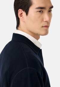 Námornícky modrý sveter s golierom, s mäkkou textúrou a viditeľnými švovými detailmi. Model má pod ním bielu košeľu.