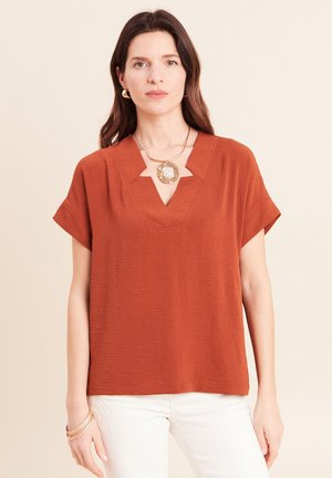 Femme portant un chemisier à manches courtes couleur rouille avec un col en V unique à encoche, un pantalon blanc, un collier pendentif en or, des boucles d'oreilles créoles et des bracelets.