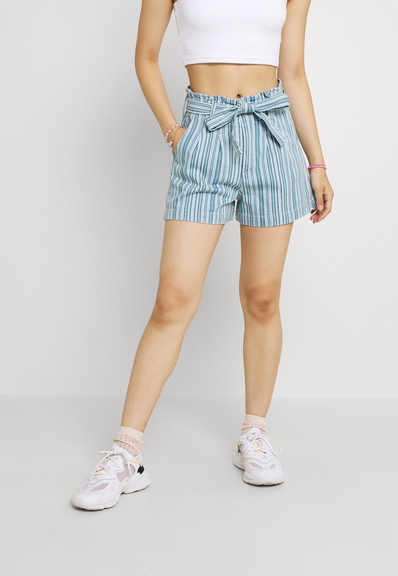 Femme portant un short rayé bleu et blanc à taille haute avec une ceinture nouée, un crop top blanc, des chaussettes roses et des baskets blanches.