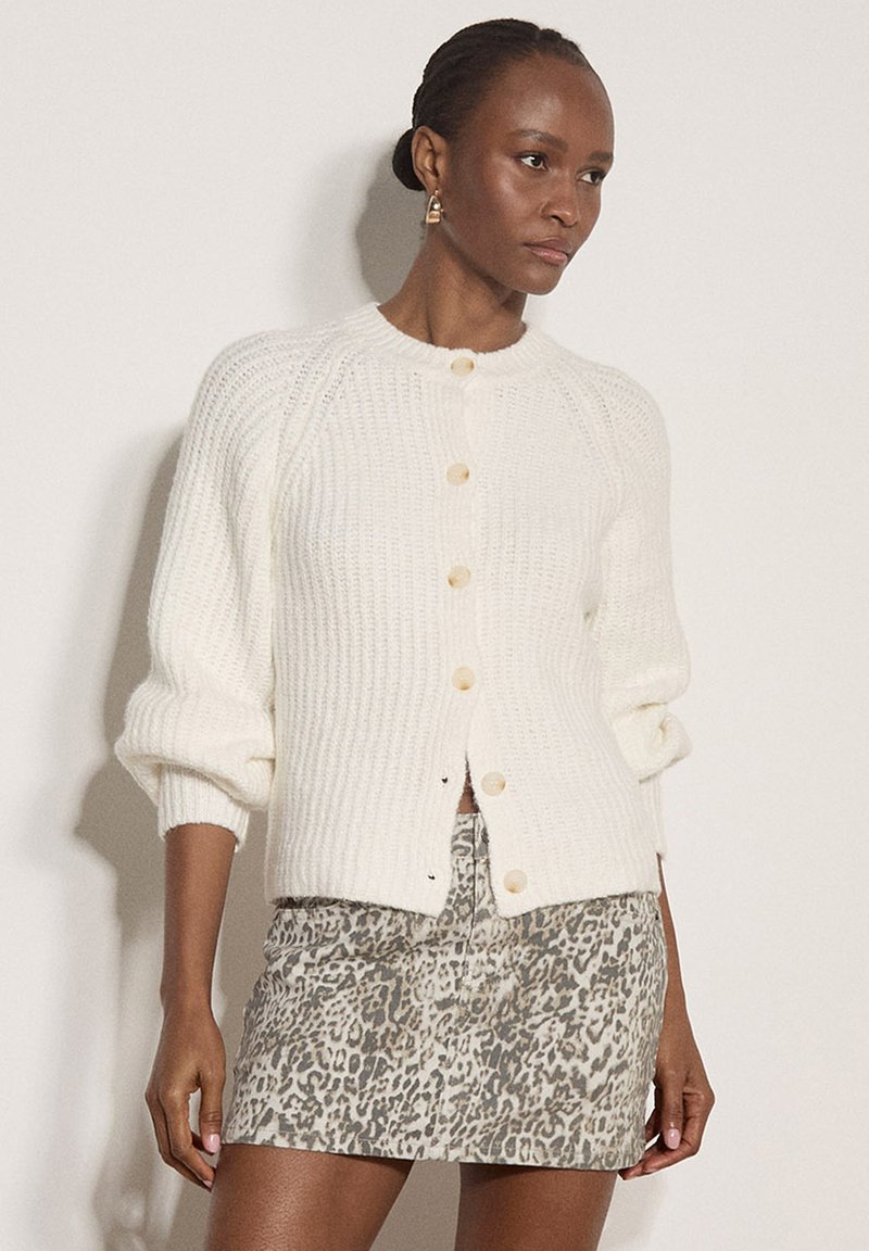 Femme portant un cardigan en tricot blanc boutonné avec les manches retroussées et une jupe courte à motif léopard, debout devant un fond uni.