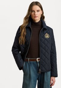 Veste matelassée noire avec fermeture éclair sur le devant, col et emblème brodé. Portée sur un pull à col roulé marron et associée à un pantalon en denim ample.