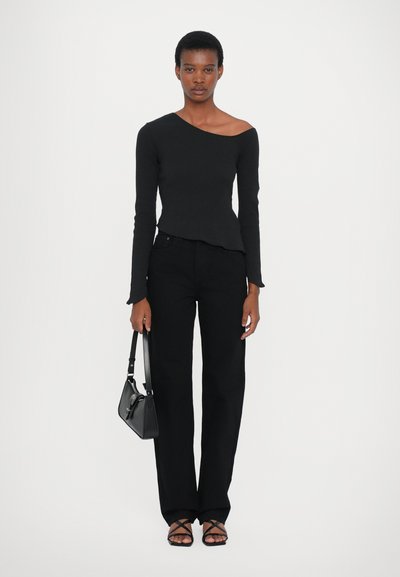 Top nero a maniche lunghe con scollo asimmetrico, abbinato a pantaloni neri a gamba dritta. Il modello tiene una piccola borsa nera e indossa sandali con cinturini.