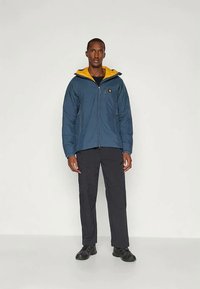 Μπλε και κίτρινη μ insulated σακάκι με κουκούλα, μπροστινό φερμουάρ και πλαϊνές τσέπες. Φορεμένο με μαύρα παντελόνια και μαύρες μπότες.