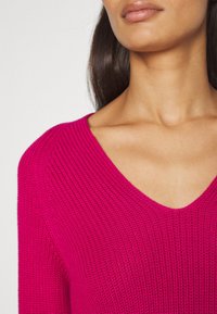 Femme portant un pull en tricot rose vif à col en V, montrant les épaules ainsi qu'une partie du cou et des lèvres sur un fond uni.