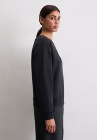 Jeune femme aux cheveux bruns dans une queue de cheval, portant un sweat-shirt à manches longues noir, se tenant de côté contre un mur blanc texturé.