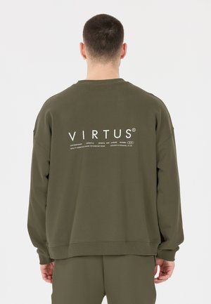 Homme aux cheveux courts portant un sweat-shirt et un pantalon vert olive, de dos, le sweat-shirt affiche "VIRTUS" en grandes lettres blanches.