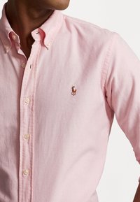Knapp-down skjorta i ljusrosa tyg, med en liten broderad hästlogotyp på vänster bröst och vita plastknappar.