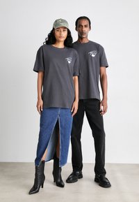 Dos modelos llevan ropa casual. Uno viste una camiseta gris y una falda de mezclilla, el otro lleva una camiseta gris a juego y pantalones negros. Botas texturizadas.
