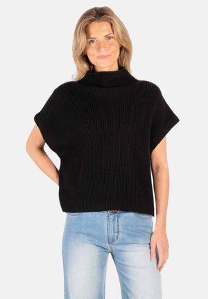 Kvinne med blondt hår iført svart turtleneck-genser med korte ermer og lyseblå jeans, står med en hånd på hoften mot hvit bakgrunn.