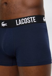 Σκούρο μπλε μποξεράκια με μαύρη ζώνη μέσης που φέρει λευκή επιγραφή "LACOSTE" και λογότυπα κροκοδείλου στη μέση και στους μηρούς ενός ατόμου.