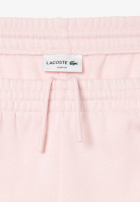 Pantalones de chándal ajustados en rosa claro con cinturilla elástica, cierre con cordón y etiqueta de marca Lacoste. Textura suave y diseño minimalista.