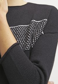 Maglione nero lavorato a maglia con un motivo bianco testurizzato sul petto, con dettagli lavorati a maglia aggiuntivi sulle maniche. Vista ravvicinata.