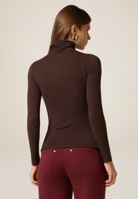 Maglione a maniche lunghe marrone con collo alto, dal design aderente e texture liscia, abbinato a pantaloni bordeaux con due tasche posteriori.