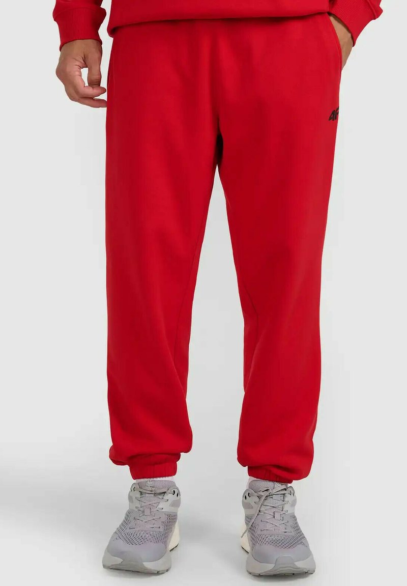 Mężczyzna ubrany w czerwone spodnie dresowe i szare buty sportowe stojący na tle jasnego, jednolitego tła.