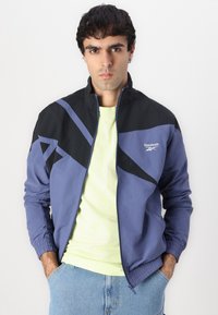 Blå og sort Reebok track jacket med geometriske mønstre, lynlås foran og elastiske manchetter, lagd over en lysegrøn t-shirt.