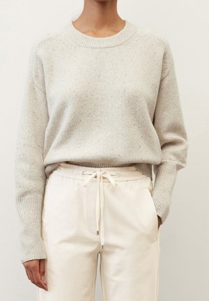 Person wearing a light beige knitted sweater tucked into off-white drawstring pants with hands relaxed by sides against a plain background.