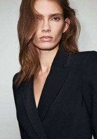 Czarny żakiet z głębokim dekoltem w kształcie V i wyraźnymi ramionami, o gładkiej teksturze. Włosy lekko potargane, otaczające twarz.