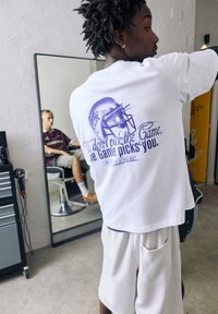 Hvid t-shirt med et lilla grafik af en fodboldhjelm og tekst, parret med lysegrå shorts. Baggrunden inkluderer et spejl og en frisørstol.