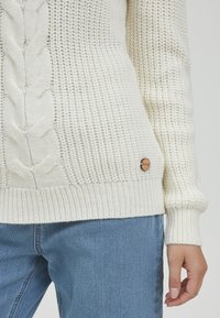 Chunky hvit strikkegenser med kabelmønster og ribbestrikkede mansjetter. Har en liten trelogo som accent på den nedre siden. Kombinert med lyseblå jeans.