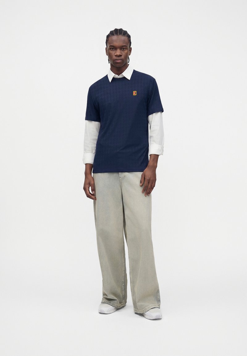 Man with braided hair stands wearing navy short-sleeve shirt over a white long-sleeve shirt, light wide-leg pants, and white sneakers.