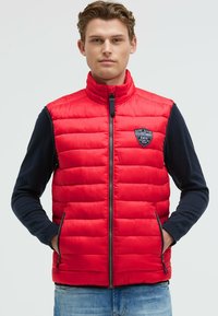 Rote gepolsterte Weste mit hohem Kragen, Reißverschluss und Logo-Patch. Mit seitlichen Taschen und glatter Stoffstruktur. Über einem dunkelblauen Langarmshirt getragen.