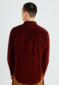 Hombre de espaldas con una camisa de manga larga de pana roja oscura y pantalones beige, contra un fondo claro sencillo.