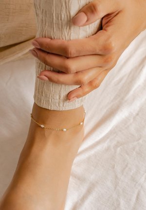 Goldene Fußkette mit abwechselnd kleinen weißen Perlen, zartes Design, kreisförmige Glieder, getragen an einem nackten Knöchel auf einem hellen, strukturierten Stoff.