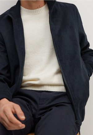 Homme portant un pull en tricot blanc sous une veste zippée bleu marine foncé et un pantalon foncé, assis avec une main reposant sur le genou.