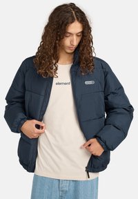 Persona con lunghi capelli ricci che indossa una giacca imbottita blu navy sopra una maglietta beige con la scritta "element" e jeans chiari a fantasia blu.