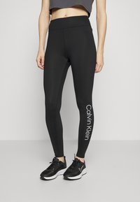 Zwarte leggings met een gladde textuur, hoge tailleband en het witte "Calvin Klein" logo dat verticaal op het linker been is gedrukt.