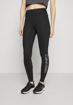 Collants noirs avec une texture lisse, taille haute et logo "Calvin Klein" imprimé verticalement sur la jambe gauche.