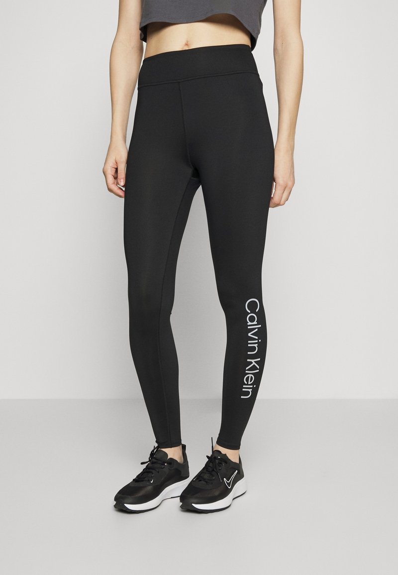 Zwarte leggings met een gladde textuur, hoge tailleband en het witte "Calvin Klein" logo dat verticaal op het linker been is gedrukt.