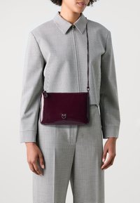 Tamnocrvena patenta kožna torba preko tijela s zatvaranjem na patentni zatvarač i reljefnim logotipom. Glatka tekstura, pravokutni oblik i podesiva traka.