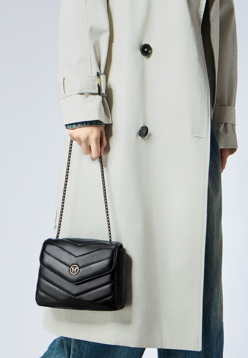 Borsa a tracolla nera imbottita con catena, dotata di chiusura a patta magnetica e accentuazione in metallo del logo, tenuta da una persona in un cappotto beige.