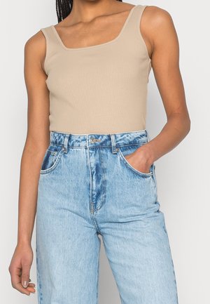 Vrouw draagt een beige geribd mouwloos topje en lichtblauwe hoge taille spijkerbroek, één hand in de zak, tegen een effen achtergrond.