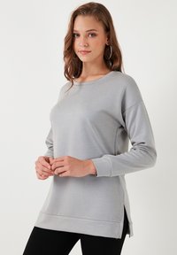 Svijetlosiva dugorukava dukserica napravljena od mekanog materijala, s okruglim izrezom, bočnim prorezima i slobodnim krojem. Istaknuti detalji uključuju šavove.