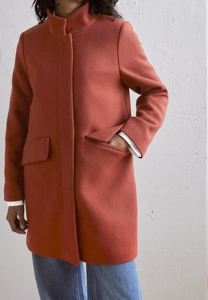 Osoba ubrana w rdzawego koloru wełniany płaszcz z klapowymi kieszeniami, stojąca z jedną ręką w kieszeni, na tle fakturowanej jasnoszarej ściany.