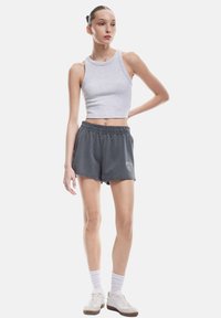 Grijze cropped tanktop en grijze sportshorts met elastische tailleband en logo. Model draagt witte sneakers en geribbelde sokken. Eenvoudig ontwerp.